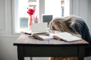 Blonde woman slumped over desk filled with books, showcasing study fatigue indoors.